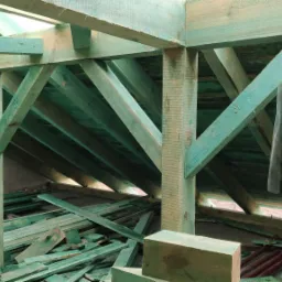 Konstrukcja więźby dachowej z impregnowanego drewna w trakcie budowy, widoczne elementy mocujące, resztki materiałów budowlanych i fragment muru.