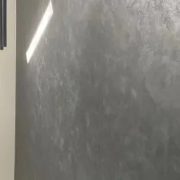 Szara ściana z efektem dekoracyjnym, oświetlona smugą światła, widoczny fragment białej listwy przypodłogowej.