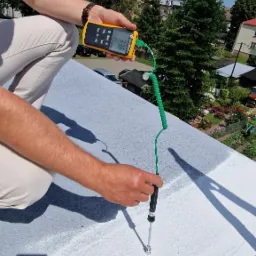 Pomiar temperatury powierzchni dachu termometrem z sondą, widoczny wynik 33.6 stopni Celsjusza, w tle zabudowa jednorodzinna i błękitne niebo z chmurami.