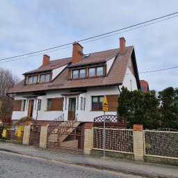 ARBAL SPÓŁKA Z OGRANICZONĄ ODPOWIEDZIALNOŚCIĄ - Dwukondygnacyjny dom z brązowym dachem i białą elewacją, widoczny z ulicy, z ogrodzeniem z cegły i metalu, w tle drzewa bez liści i pochmurne niebo.