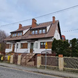 Dwukondygnacyjny dom z brązowym dachem i białą elewacją, widoczny z ulicy, z ogrodzeniem z cegły i metalu, w tle drzewa bez liści i pochmurne niebo.