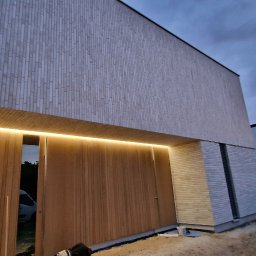 ARBAL SPÓŁKA Z OGRANICZONĄ ODPOWIEDZIALNOŚCIĄ - Nowoczesny dom z elewacją z jasnego drewna i cegły, podświetlony ledami nad wejściem, widok na zachodzące słońce w tle.