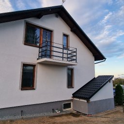 ARBAL SPÓŁKA Z OGRANICZONĄ ODPOWIEDZIALNOŚCIĄ - Boczny widok na dom po renowacji elewacji, z balkonem i oknami w drewnianych ramach, z widocznym fragmentem ogrodu i sąsiednich budynków w tle.