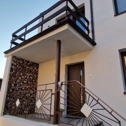 ARBAL SPÓŁKA Z OGRANICZONĄ ODPOWIEDZIALNOŚCIĄ - Dwupiętrowy dom z białymi ścianami, balkonem z kamienną mozaiką i metalową balustradą ze wzorem słońca, widok z dołu w słoneczny dzień.