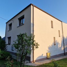 ARBAL SPÓŁKA Z OGRANICZONĄ ODPOWIEDZIALNOŚCIĄ - Dwukondygnacyjny dom z jasną elewacją w promieniach popołudniowego słońca, widoczny narożnik budynku z brązowymi oknami i rynnami, na pierwszym planie zielona roślinność.