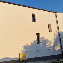 Jasna elewacja budynku z trzema wąskimi oknami, oświetlona popołudniowym słońcem, z widocznym żółtym skrzynką na listy i cieniem drzewa.