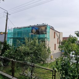 ARBAL SPÓŁKA Z OGRANICZONĄ ODPOWIEDZIALNOŚCIĄ - Dom w trakcie prac elewacyjnych, rusztowanie z siatką ochronną, widoczny baner reklamowy firmy budowlanej, częściowo otynkowana ściana, drabina oparta o dach.