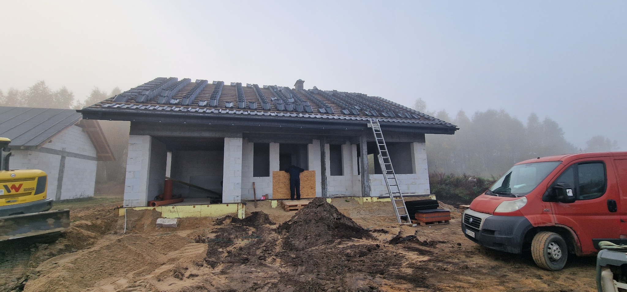 Budowa domu jednorodzinnego w trakcie realizacji: częściowo ułożone dachówki, ściany z bloczków, widoczna drabina i czerwony samochód dostawczy na zewnątrz w pochmurny dzień.