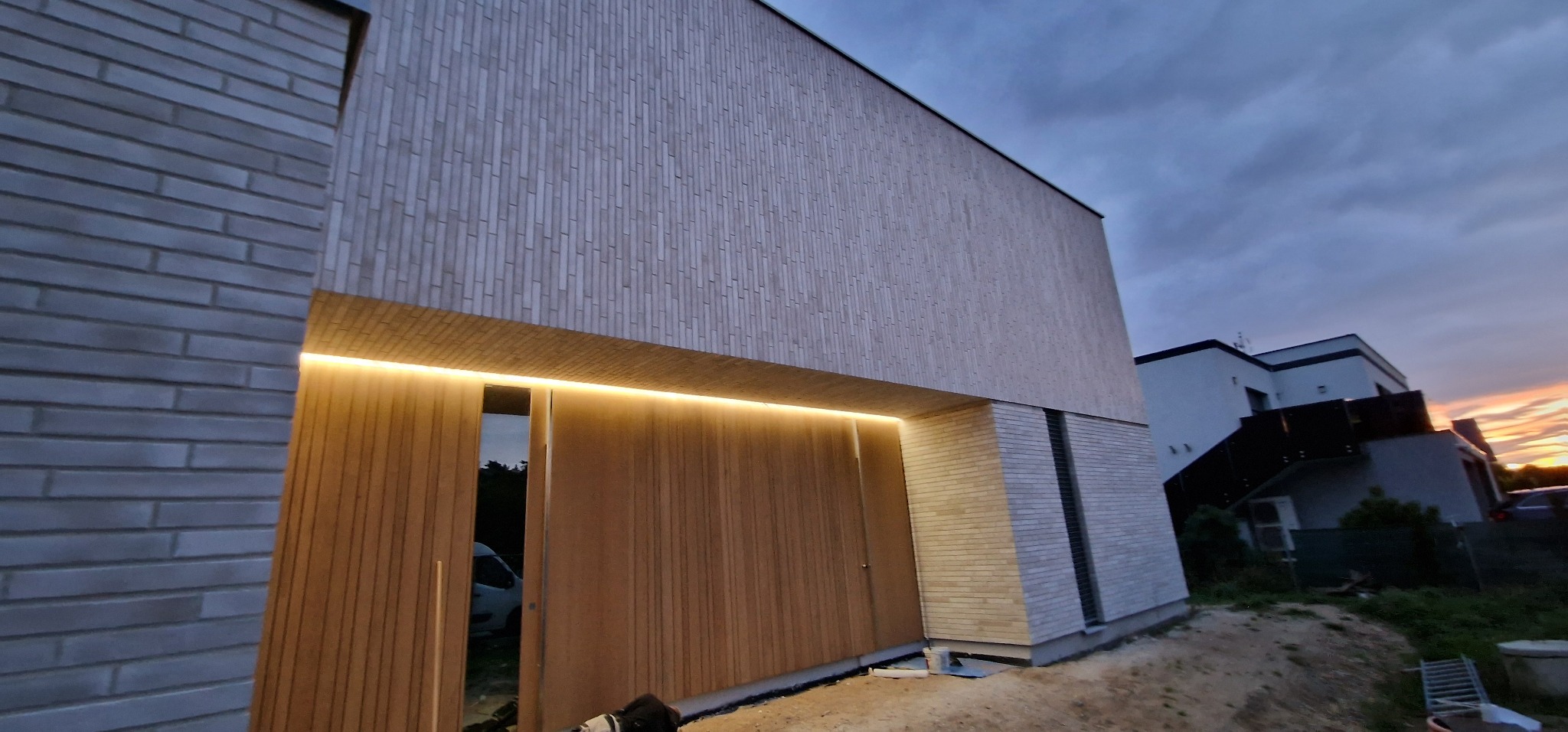 Nowoczesny dom z elewacją z jasnego drewna i cegły, podświetlony ledami nad wejściem, widok na zachodzące słońce w tle.