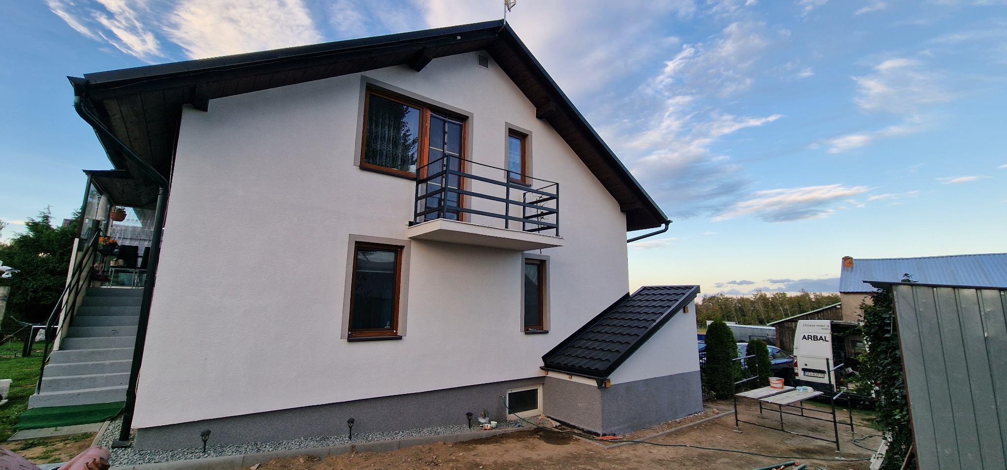 Boczny widok na dom po renowacji elewacji, z balkonem i oknami w drewnianych ramach, z widocznym fragmentem ogrodu i sąsiednich budynków w tle.