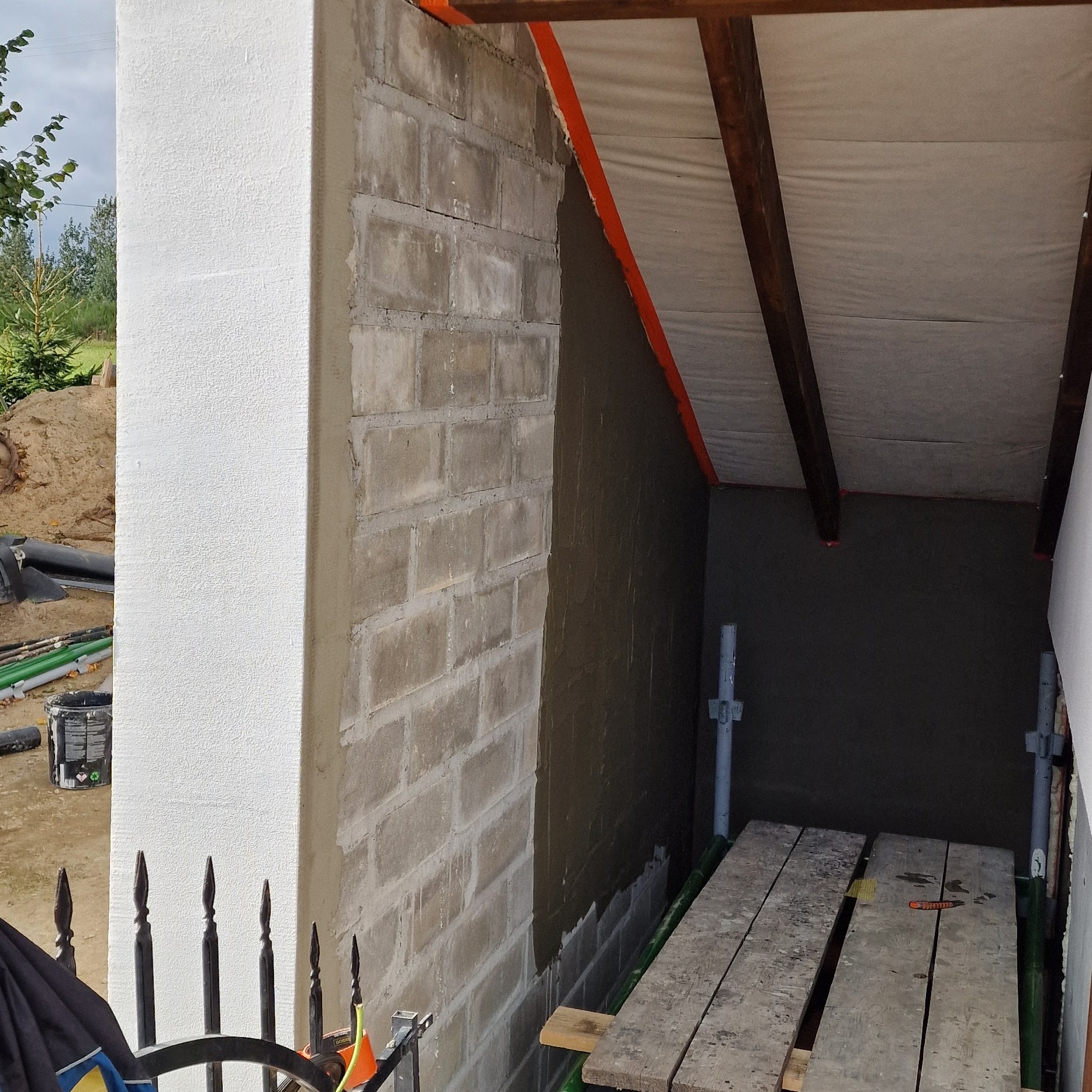 Fragment ściany budynku w trakcie prac wykończeniowych: otynkowany i pomalowany fragment elewacji kontrastuje z surowym murem z bloczków i ocieplonym fragmentem pod dachem, widoczne rusztowanie.