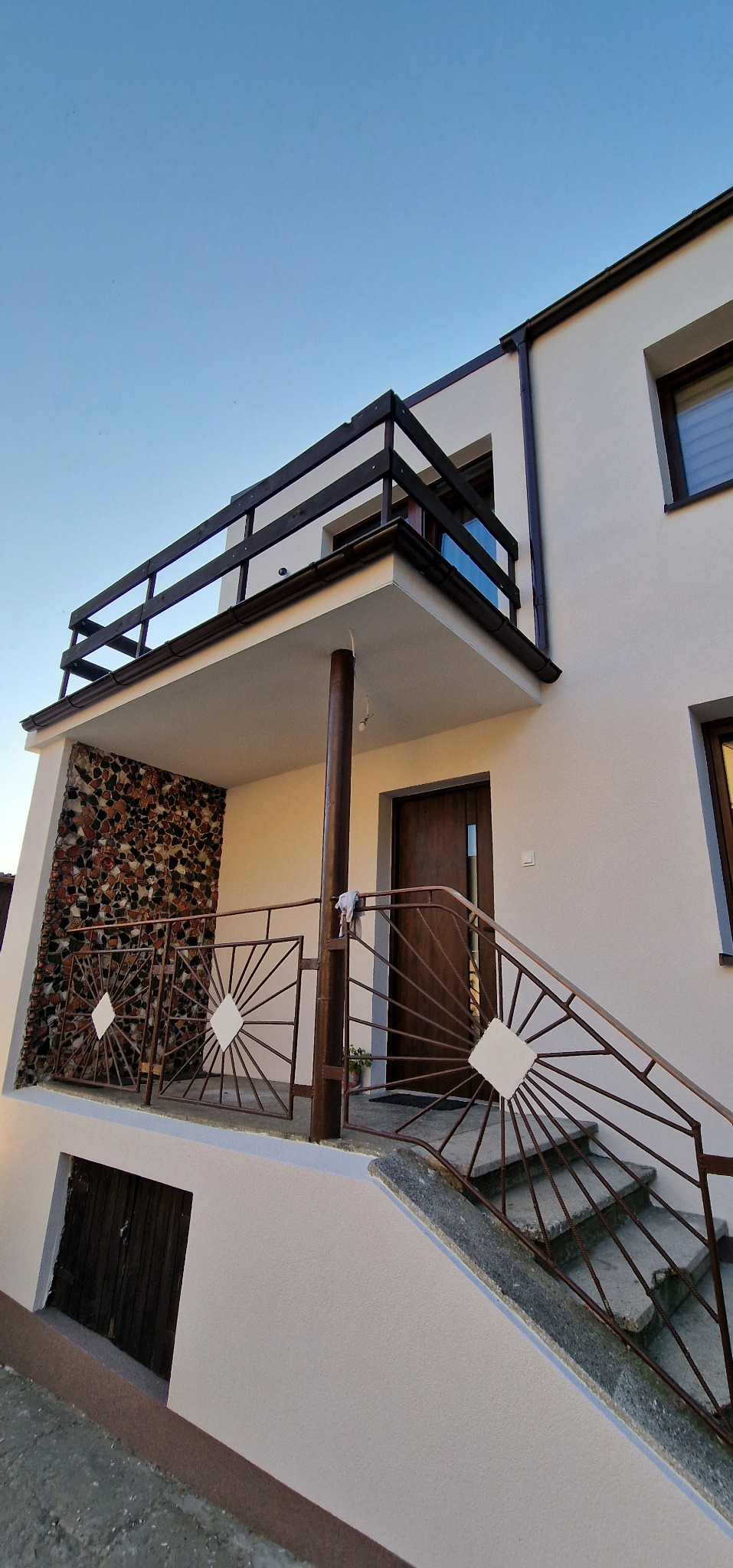 Dwupiętrowy dom z białymi ścianami, balkonem z kamienną mozaiką i metalową balustradą ze wzorem słońca, widok z dołu w słoneczny dzień.