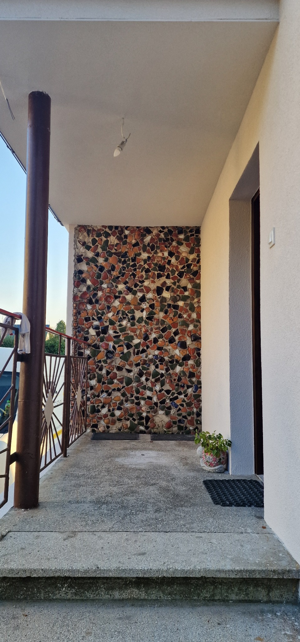 Balkon z elewacją wykończoną mozaiką z potłuczonych naczyń w odcieniach brązu, beżu i zieleni, z metalową balustradą i doniczką z rośliną.