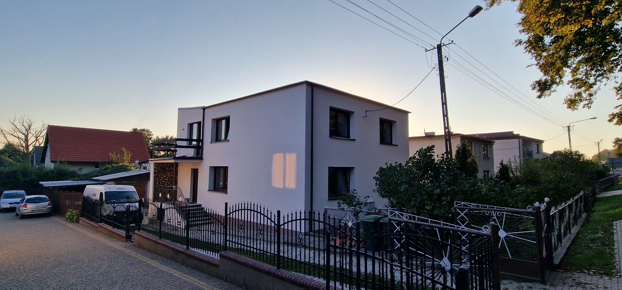 Dwukondygnacyjny dom jednorodzinny z białymi ścianami i ciemnymi ramami okiennymi, widoczny z perspektywy ulicy z metalowym ogrodzeniem i zaparkowanymi samochodami w tle, oświetlony popołudniowym...