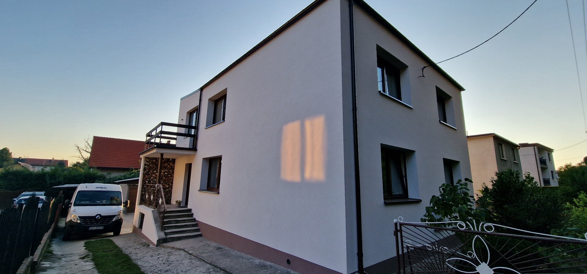 Dwupiętrowy dom z jasną elewacją i ciemnymi obramowaniami okien, widok z boku, z fragmentem ogrodzenia i zaparkowanym samochodem dostawczym.