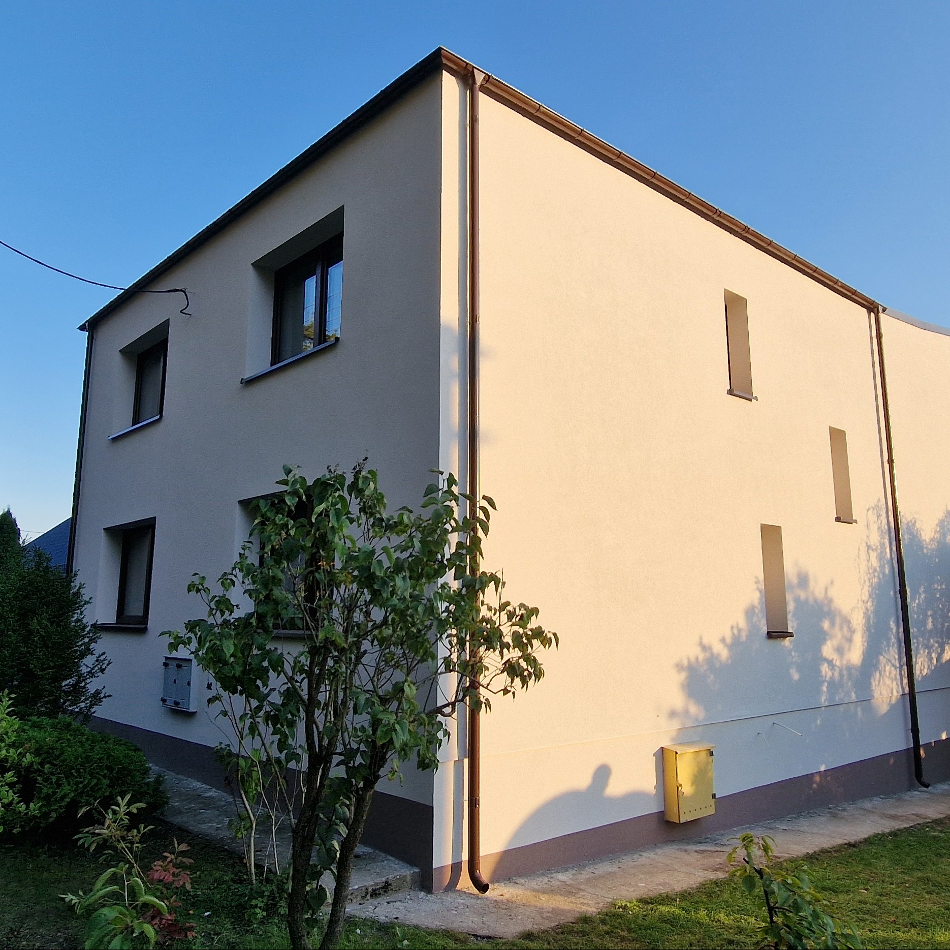 Dwukondygnacyjny dom z jasną elewacją w promieniach popołudniowego słońca, widoczny narożnik budynku z brązowymi oknami i rynnami, na pierwszym planie zielona roślinność.