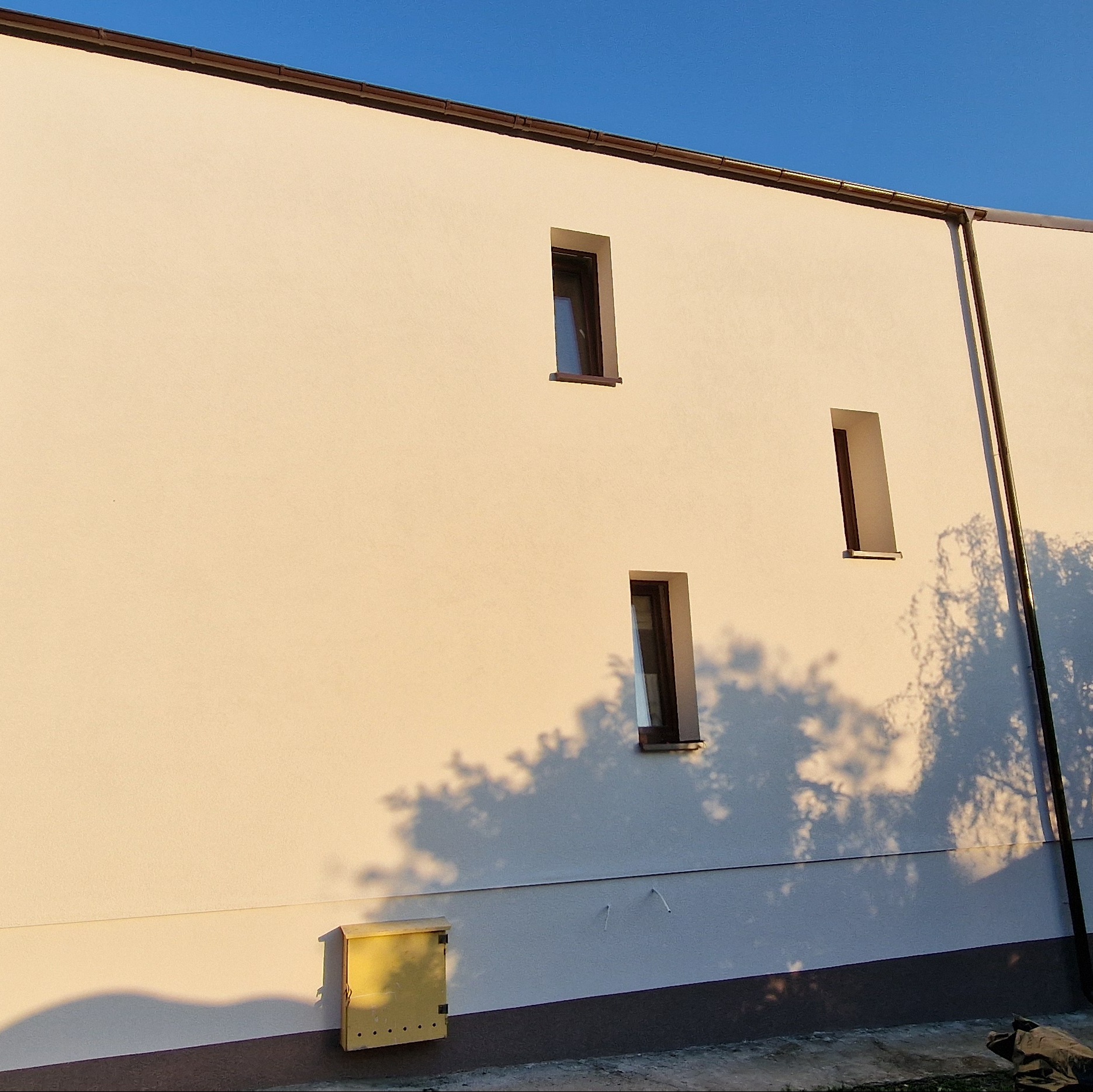 Jasna elewacja budynku z trzema wąskimi oknami, oświetlona popołudniowym słońcem, z widocznym żółtym skrzynką na listy i cieniem drzewa.