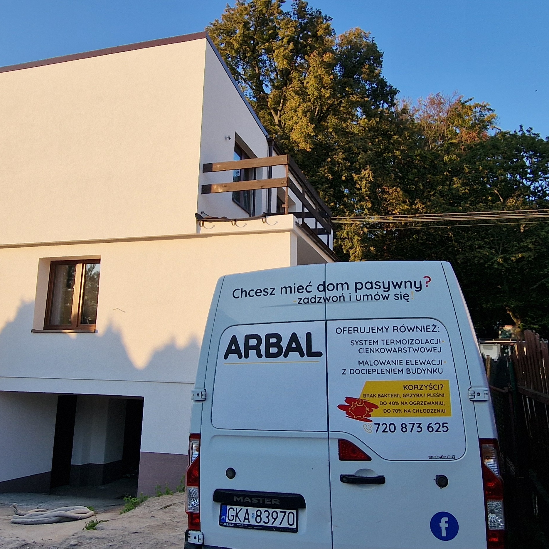Jasna elewacja domu z balkonem, częściowo zasłonięta przez firmowy samochód dostawczy z reklamą usług termoizolacyjnych i numerem telefonu.