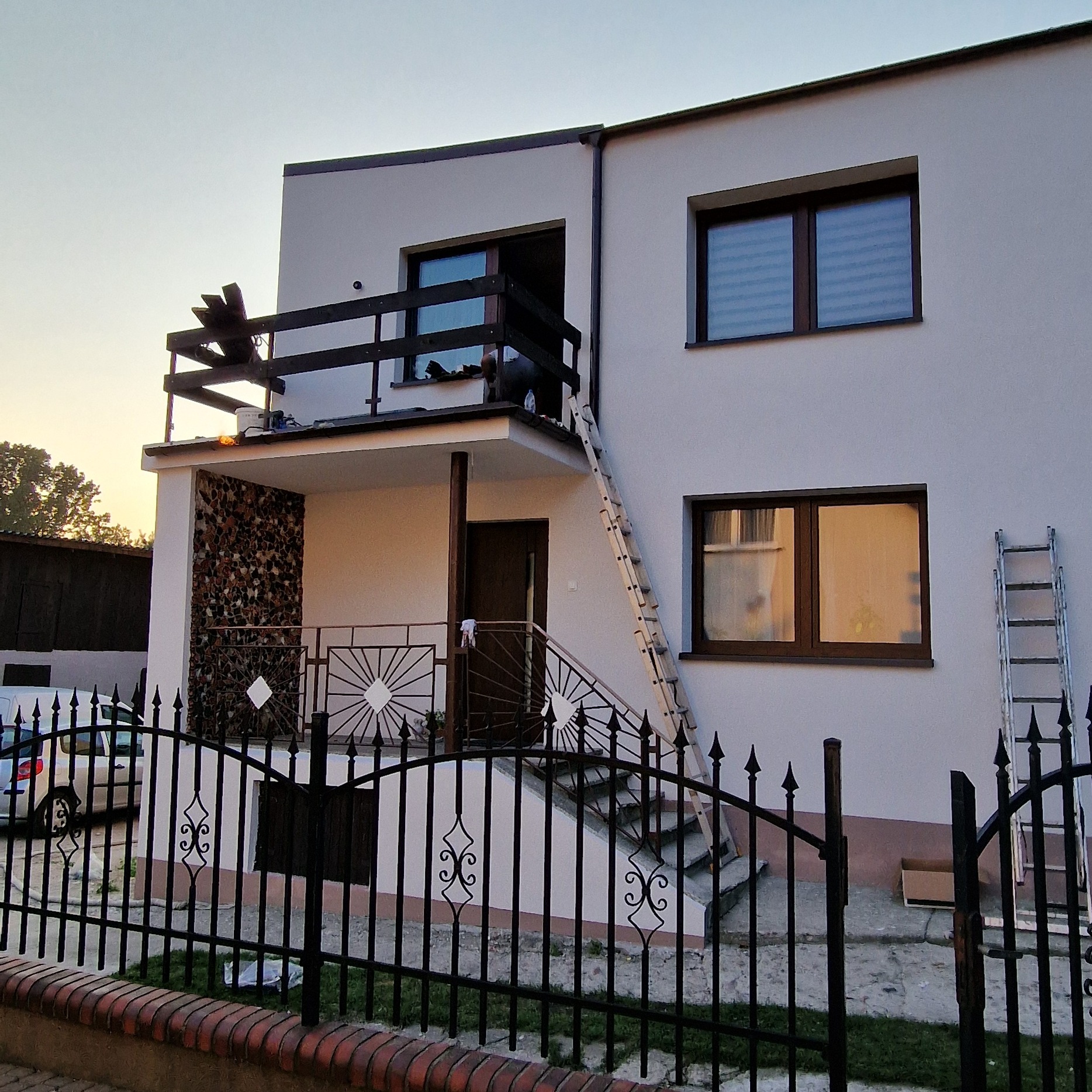 Dwupiętrowy dom z jasną elewacją w trakcie malowania, widoczne drabiny, balkon ze zdobieniami z kamienia, czarne metalowe ogrodzenie z ceglaną podmurówką.