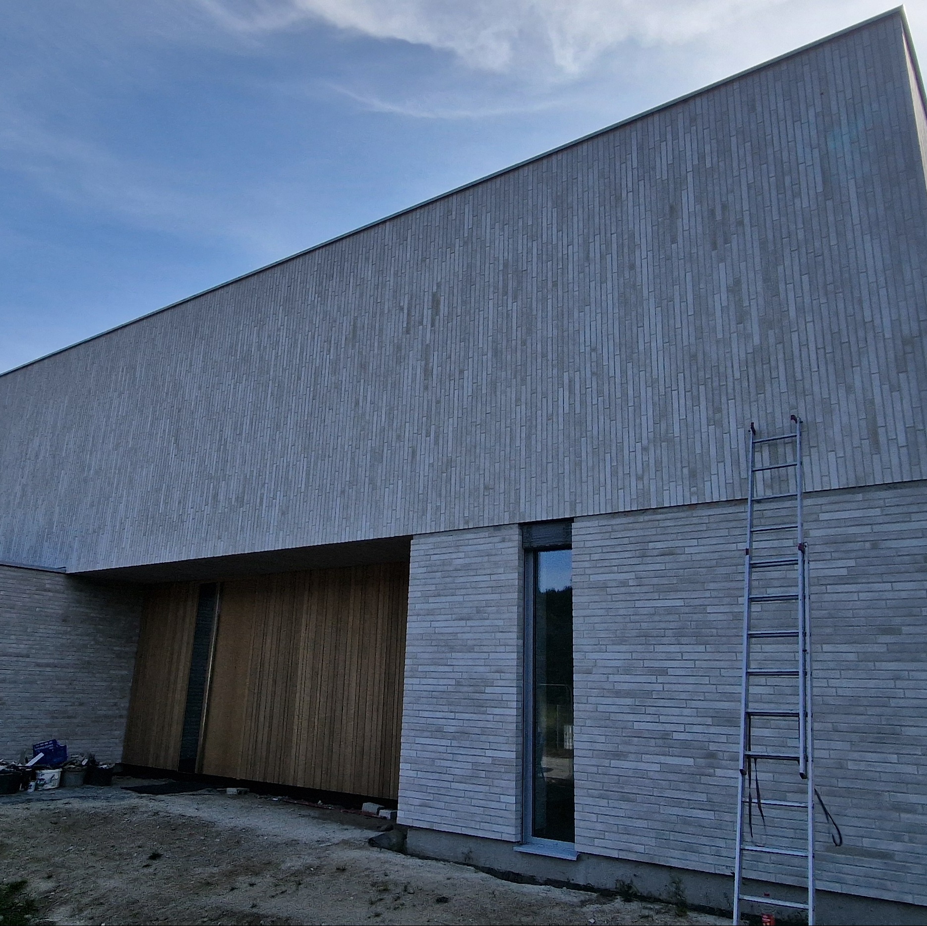 Nowoczesny budynek z szarej cegły klinkierowej, drewniane panele, aluminiowa drabina oparta o ścianę, widoczne pojemniki z zaprawą