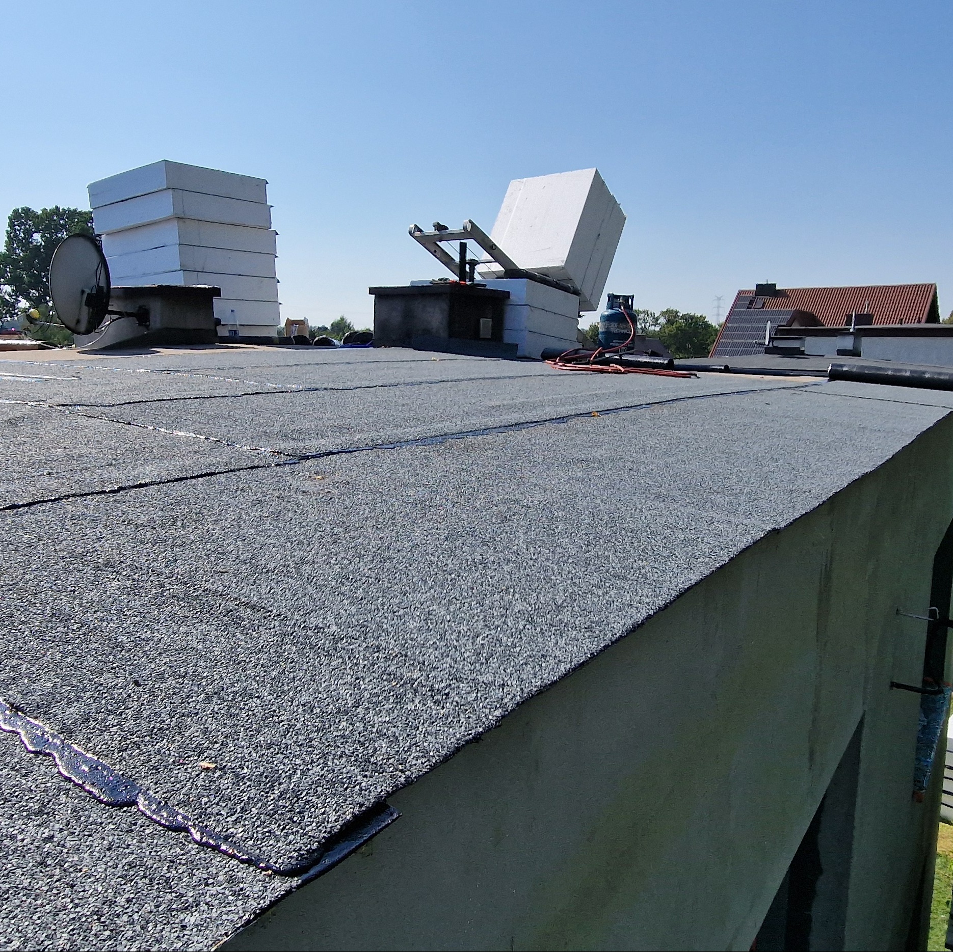 Widok na płaski dach pokryty papą z widocznymi łączeniami, na którym znajdują się stosy styropianu, antena satelitarna i butla gazowa z palnikiem dekarskim.