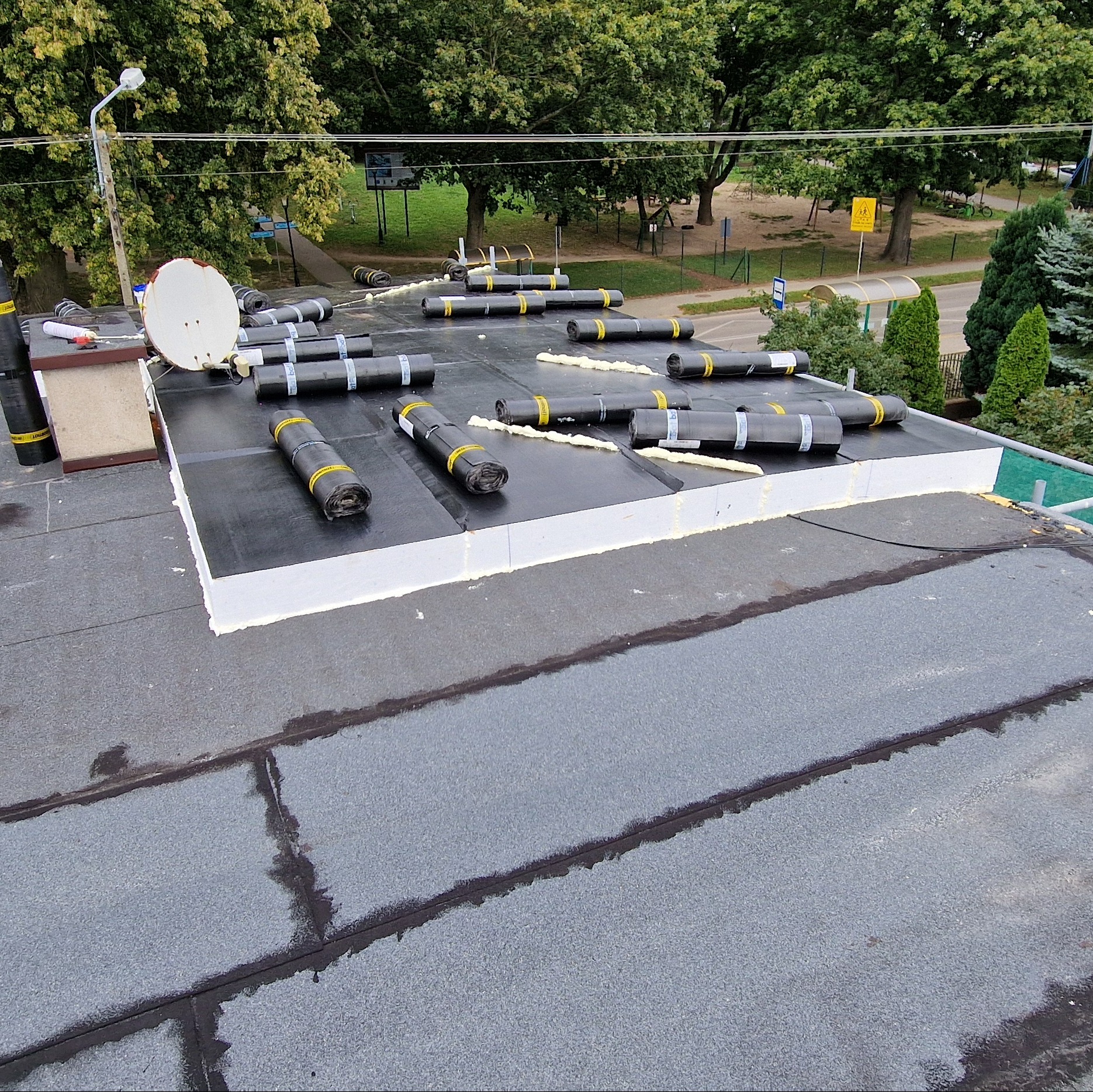 Czarny dach płaski z rolkami papy, pianką uszczelniającą i anteną satelitarną w tle parku miejskiego.