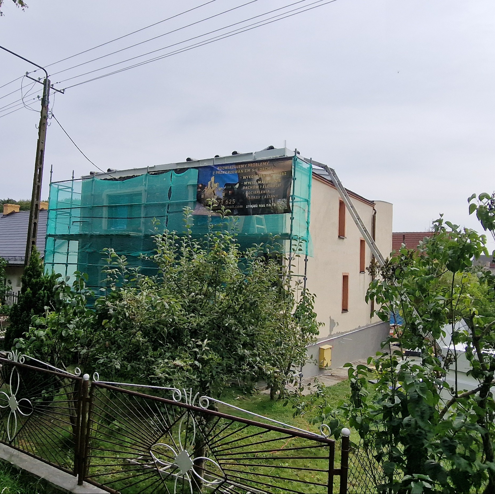 Dom w trakcie prac elewacyjnych, rusztowanie z siatką ochronną, widoczny baner reklamowy firmy budowlanej, częściowo otynkowana ściana, drabina oparta o dach.