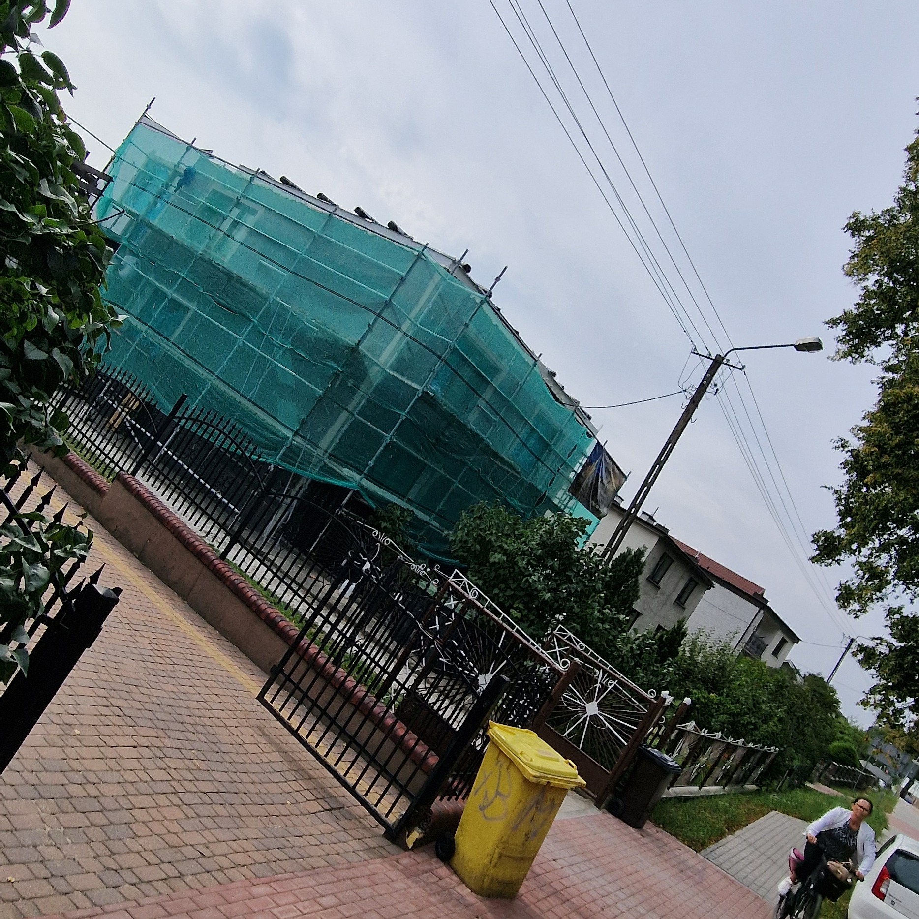 Dom w trakcie prac elewacyjnych, osłonięty zieloną siatką rusztowania, widok z chodnika z żółtym pojemnikiem na śmieci i rowerzystką w tle.