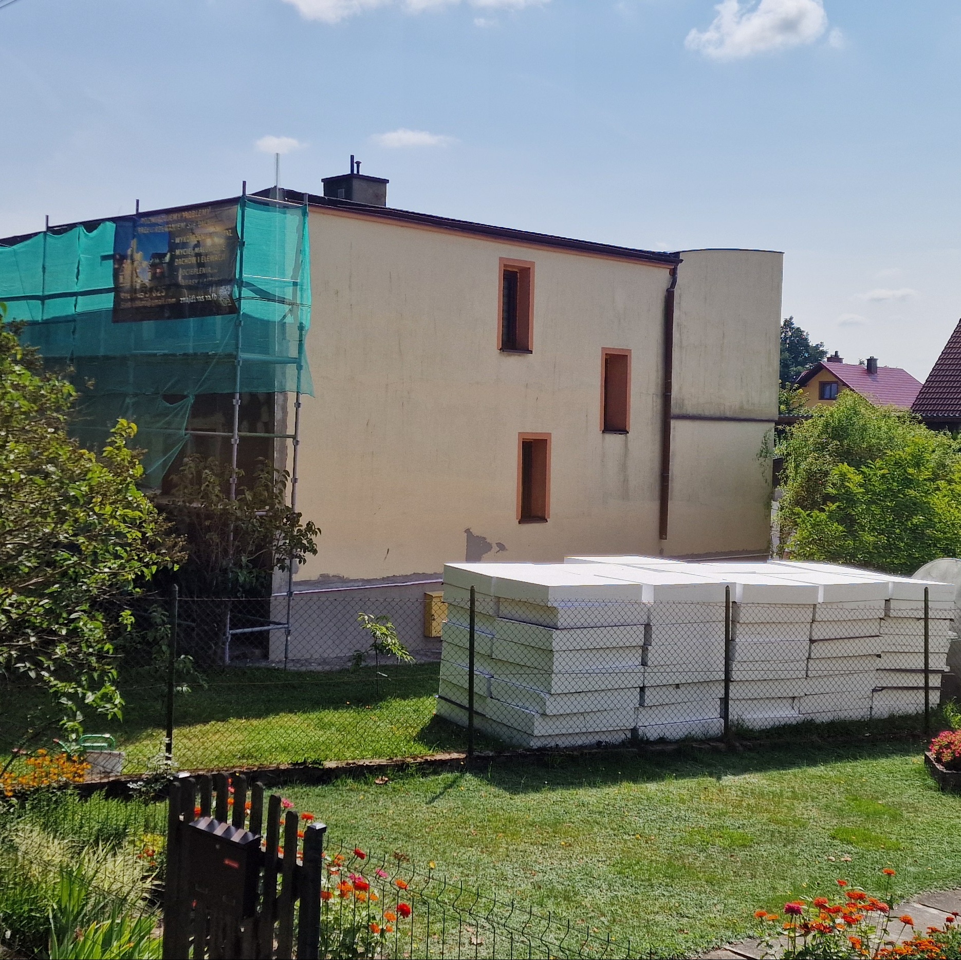 Dwupiętrowy dom z beżową elewacją w trakcie ocieplania, widoczne rusztowania z siatką ochronną oraz stos białego styropianu na trawniku.