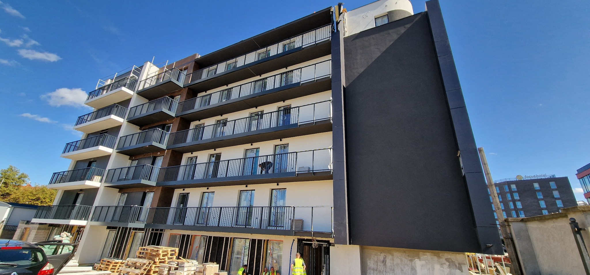 Wielopiętrowy budynek mieszkalny z balkonami, częściowo wykończony, z widocznymi pracownikami w kamizelkach odblaskowych i materiałami budowlanymi u podstawy, na tle błękitnego nieba.