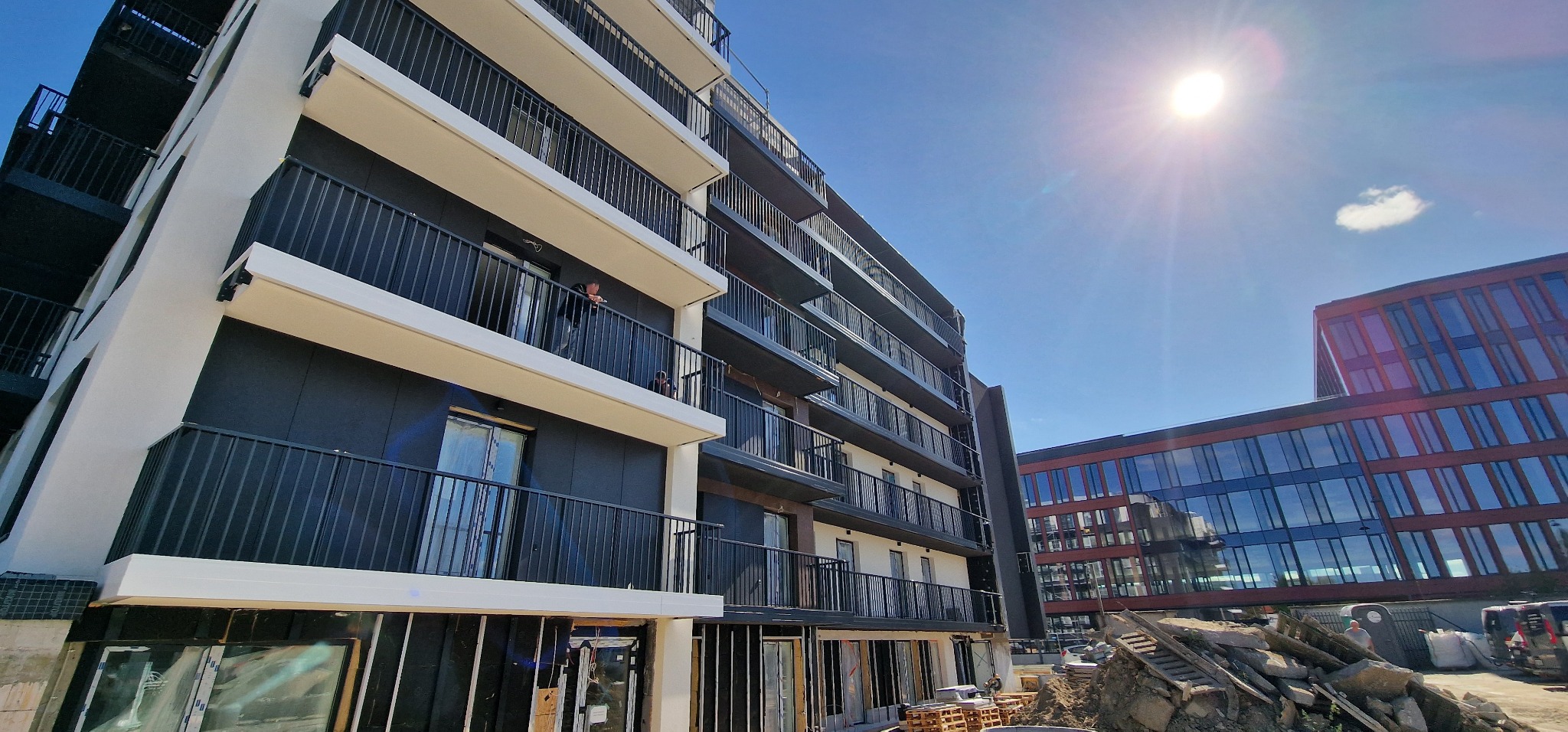 Nowoczesny budynek wielorodzinny z czarną elewacją i białymi elementami wykończeniowymi balkonów w trakcie wykańczania, widoczne rusztowania i materiały budowlane, jasne słońce na błękitnym niebie.