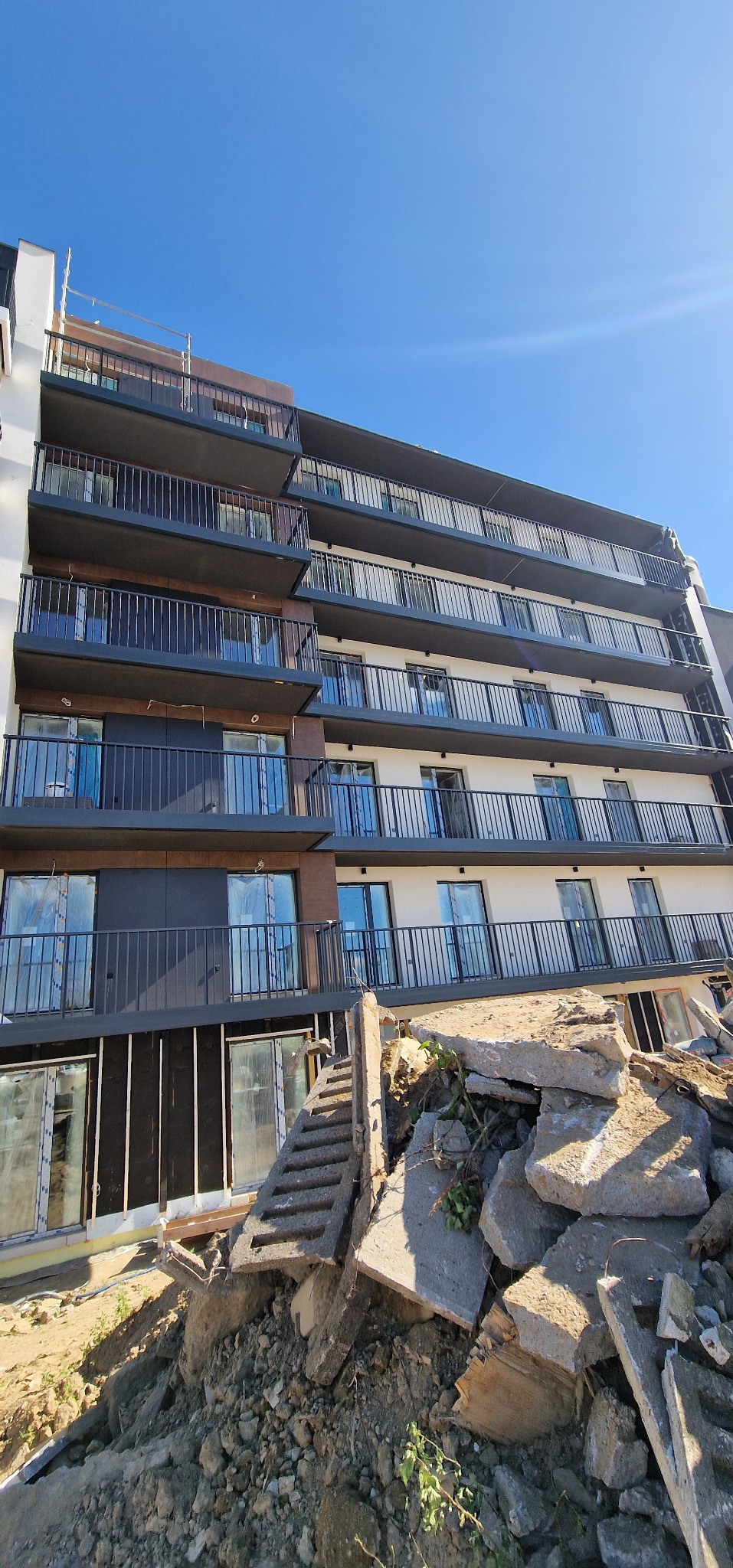 Wielopiętrowy budynek z balkonami, częściowo ocieplony, z gruzem i ziemią u podstawy, wskazującymi na trwające prace budowlane.
