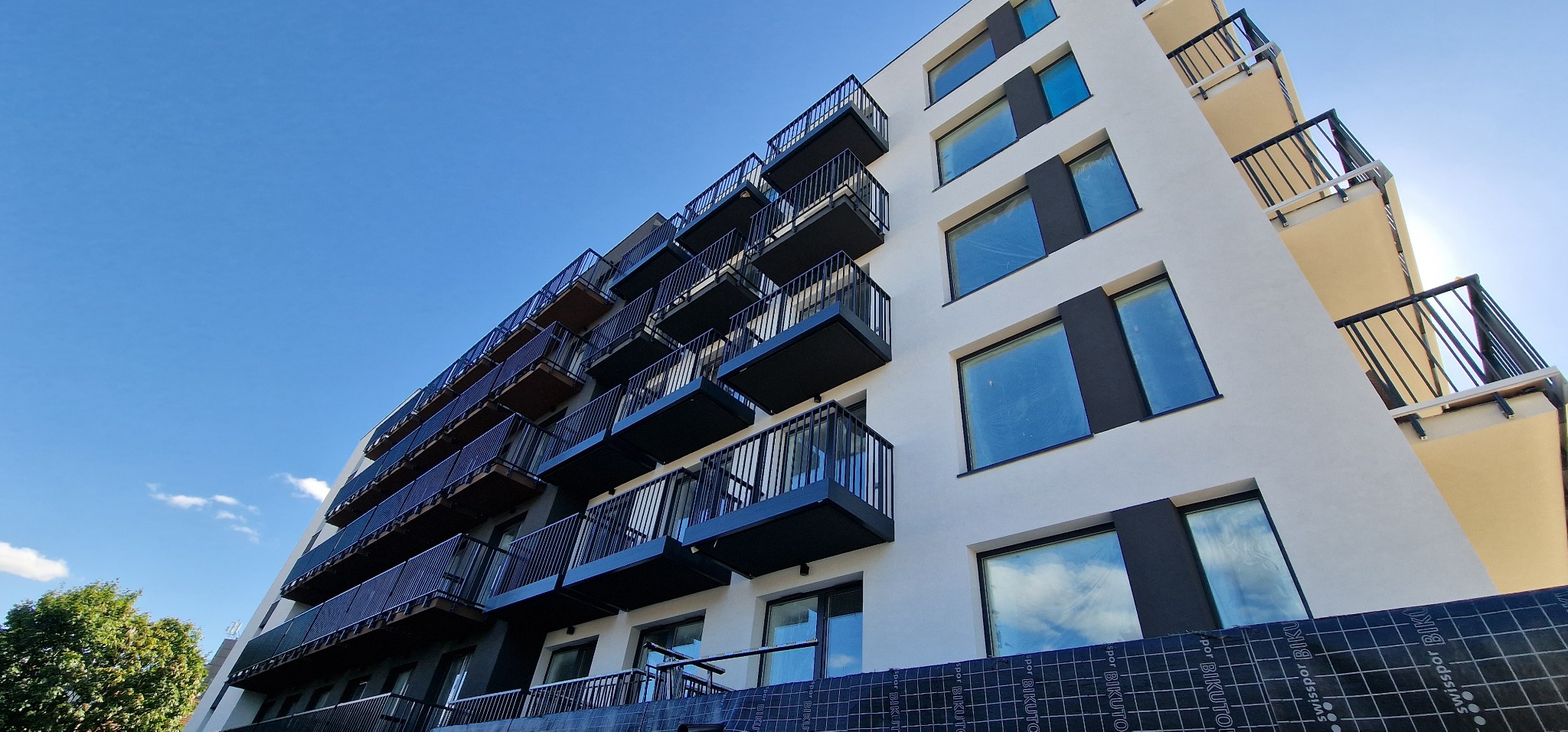 Nowoczesny budynek wielorodzinny z białymi ścianami, czarnymi obramowaniami okien i balkonami z czarnymi balustradami, widok z dołu na tle błękitnego nieba, zabezpieczenia okien folią, widoczna...