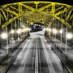 Ż&oacute;łta, stalowa konstrukcja mostu oświetlona lampami ulicznymi nocą, z widokiem na tory tramwajowe i jezdnię w odcieniach szarości, z logo URBAN GEO w dolnej części kadru.