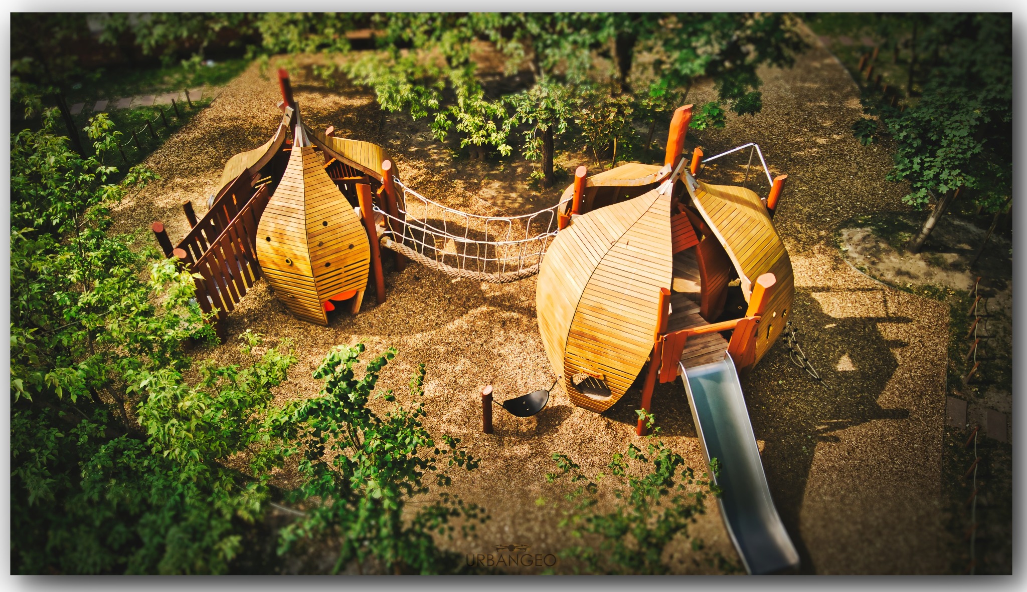 Widok z góry na drewniany plac zabaw w kształcie dwóch domków połączonych linowym mostem, z metalową zjeżdżalnią i siedziskiem na sprężynie, otoczony zielenią i korą drzewną.