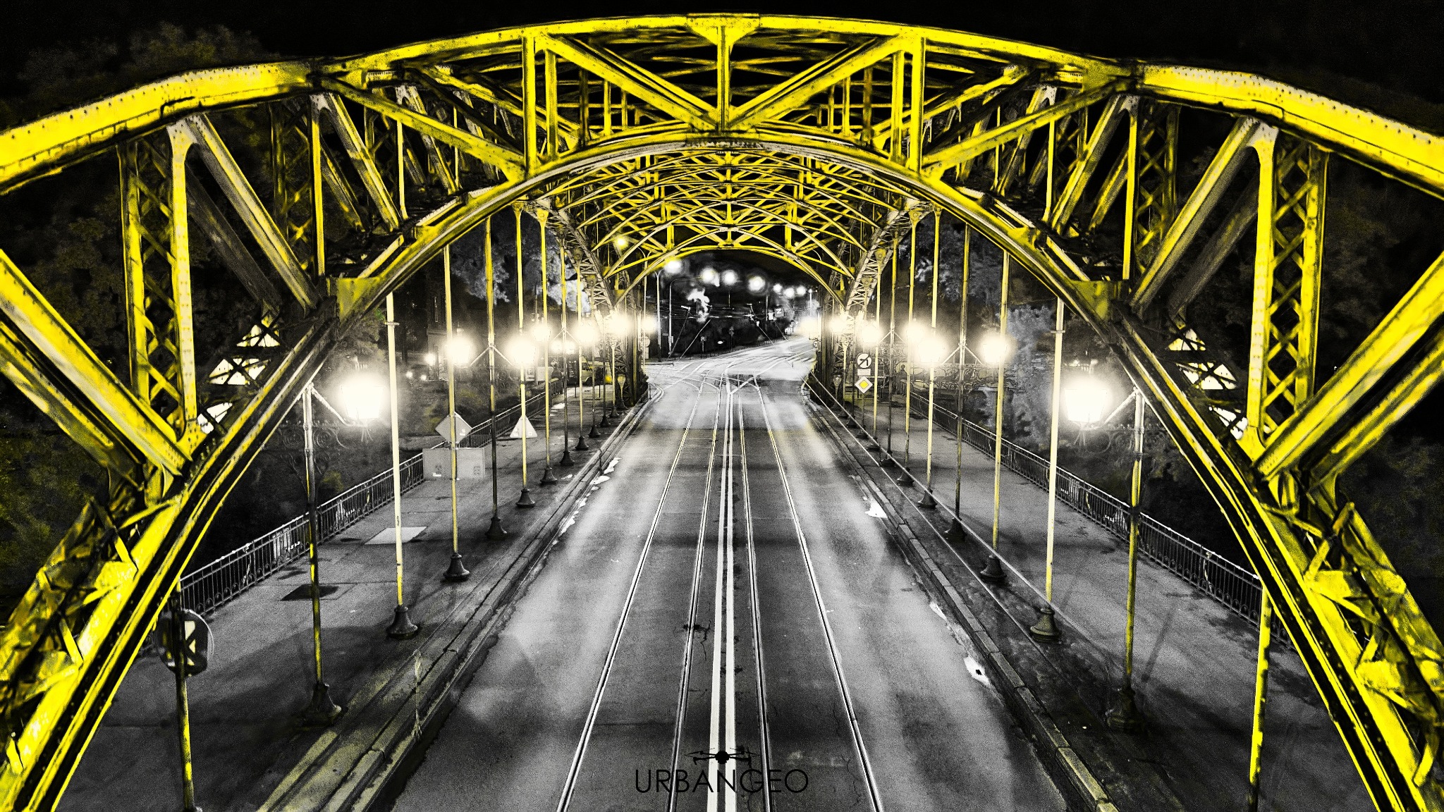 Żółta, stalowa konstrukcja mostu oświetlona lampami ulicznymi nocą, z widokiem na tory tramwajowe i jezdnię w odcieniach szarości, z logo URBAN GEO w dolnej części kadru.