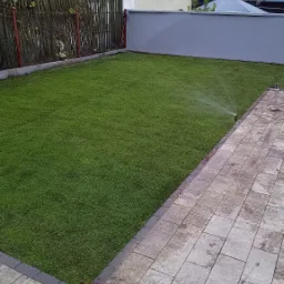 Nawodniony trawnik z rolki przylegający do brukowanego tarasu, widoczny system odwadniający, w tle ogrodzenie i dźwigi portowe.