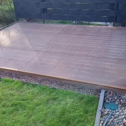 Nowoczesny taras z deski kompozytowej w odcieniach brązu, otoczony kamieniami i trawnikiem, z ciemnym, ażurowym ogrodzeniem w tle.