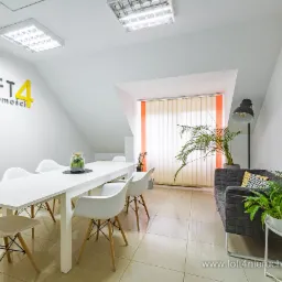 Jasne biuro nieruchomości z białym stołem konferencyjnym, krzesłami, szarą sofą, roślinami doniczkowymi i logo firmy 'LOFT4 nieruchomości' na ścianie.