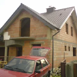 Surowy budynek jednorodzinny w trakcie budowy, z widocznymi czerwonymi cegłami, brakiem okien i otwartym bagażnikiem czerwonego samochodu osobowego na pierwszym planie.