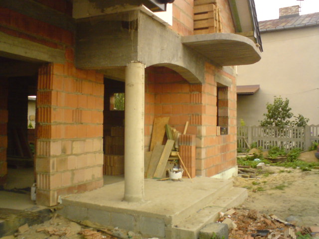 Surowy stan domu w trakcie budowy: ściany z czerwonej cegły kratówki, betonowe nadproża, kolumna podtrzymująca i widoczne deski oraz gruz budowlany na zewnątrz.