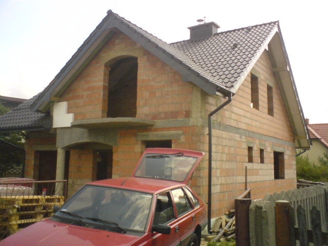 Surowy budynek jednorodzinny w trakcie budowy, z widocznymi czerwonymi cegłami, brakiem okien i otwartym bagażnikiem czerwonego samochodu osobowego na pierwszym planie.