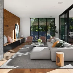 Jasny salon z drewnianą podłogą, szarą ścianą z mozaiki przy kominku, drewnianą ścianą z półką, szarą sofą narożną i dużymi oknami przesuwnymi wychodzącymi na taras z grillem.