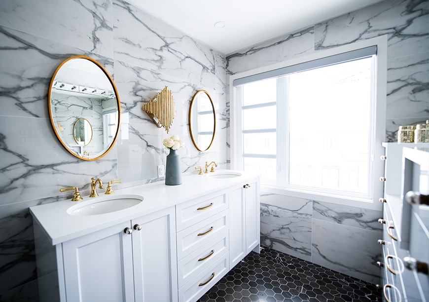 Elegancka łazienka z marmurowymi płytkami, dwiema owalnymi lustrami w złotych ramach, białymi szafkami z złotymi uchwytami, czarną mozaiką na podłodze i dekoracyjną lampą ścienną w kształcie...