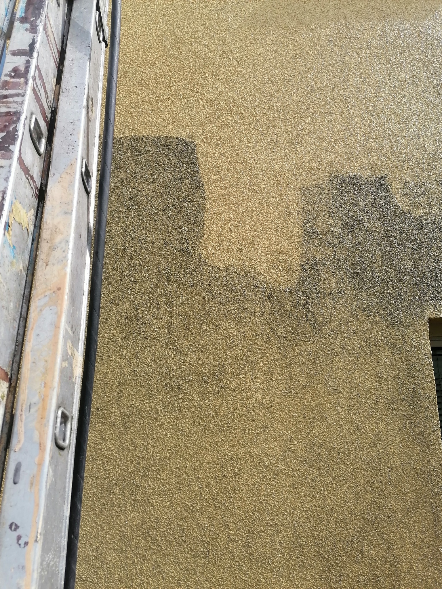 Fragment elewacji budynku z żółtym, ziarnistym tynkiem mineralnym, częściowo pokrytym ciemniejszą warstwą, z widoczną drabinką malarską z boku.