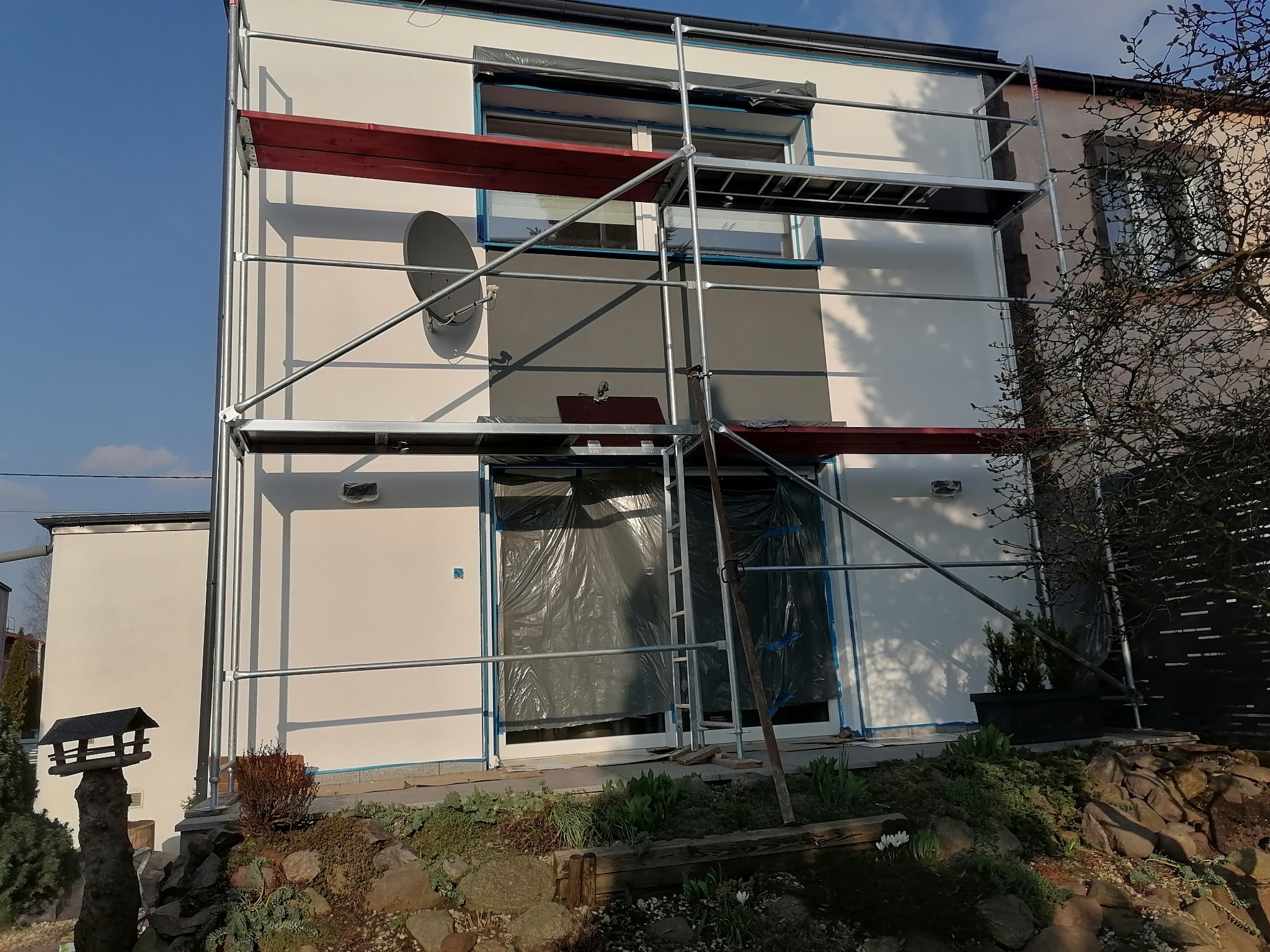 Elewacja domu w trakcie odświeżania, zabezpieczone okna folią, widoczne rusztowanie i fragment pomalowanej ściany w szarym kolorze.