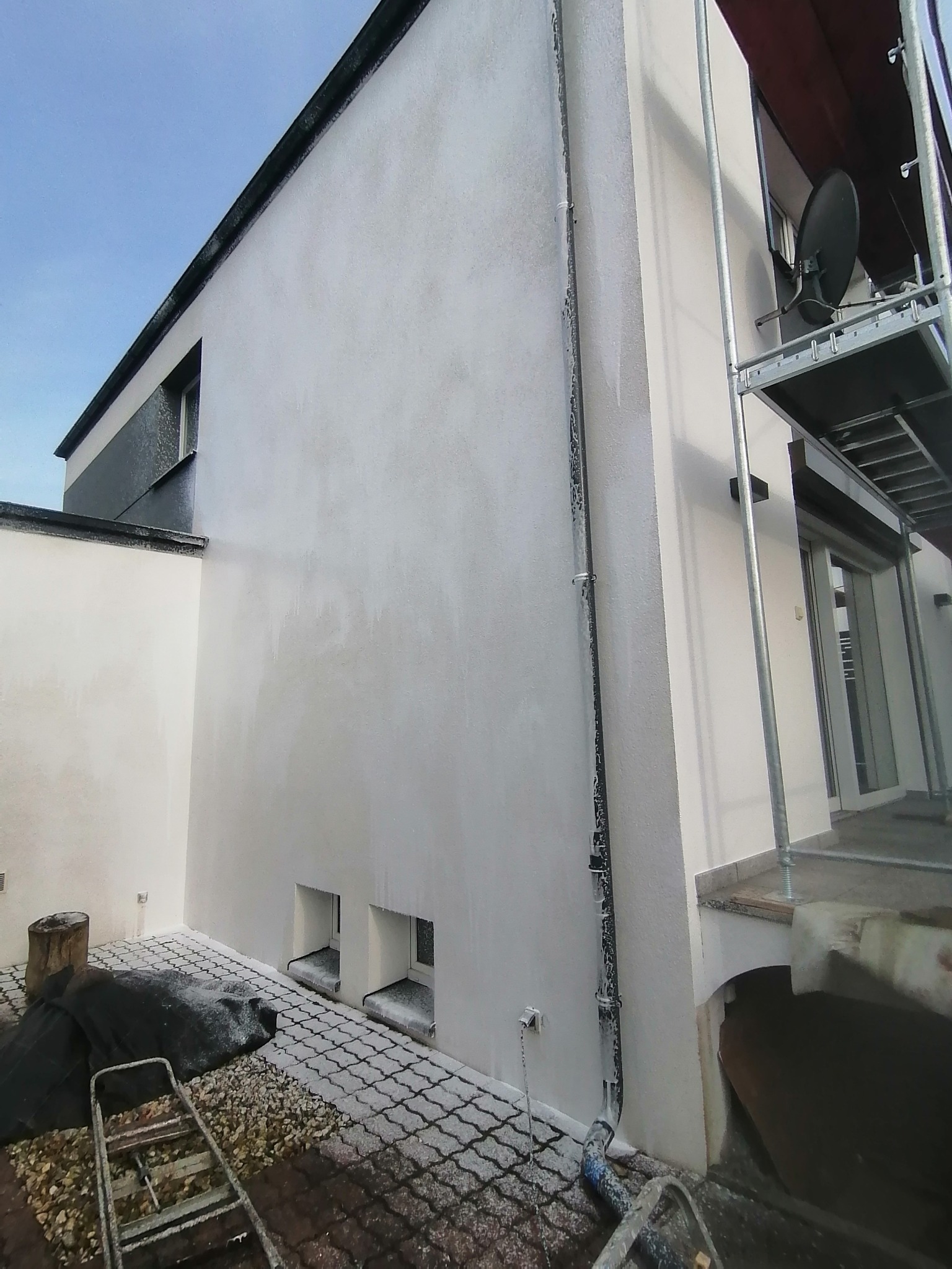 Świeżo pomalowana, biała elewacja domu jednorodzinnego, widoczna z dołu, z elementami rusztowania, rynną i fragmentem kostki brukowej pokrytej resztkami farby.