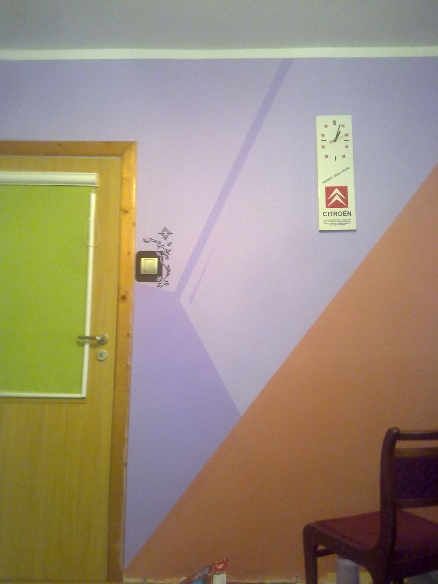 Pokój z fioletową ścianą ozdobioną geometrycznym wzorem w kolorze pomarańczowym, zegarem reklamowym Citroen, naklejką kwiatową przy włączniku światła i drewnianą ramą drzwi z zieloną roletą.