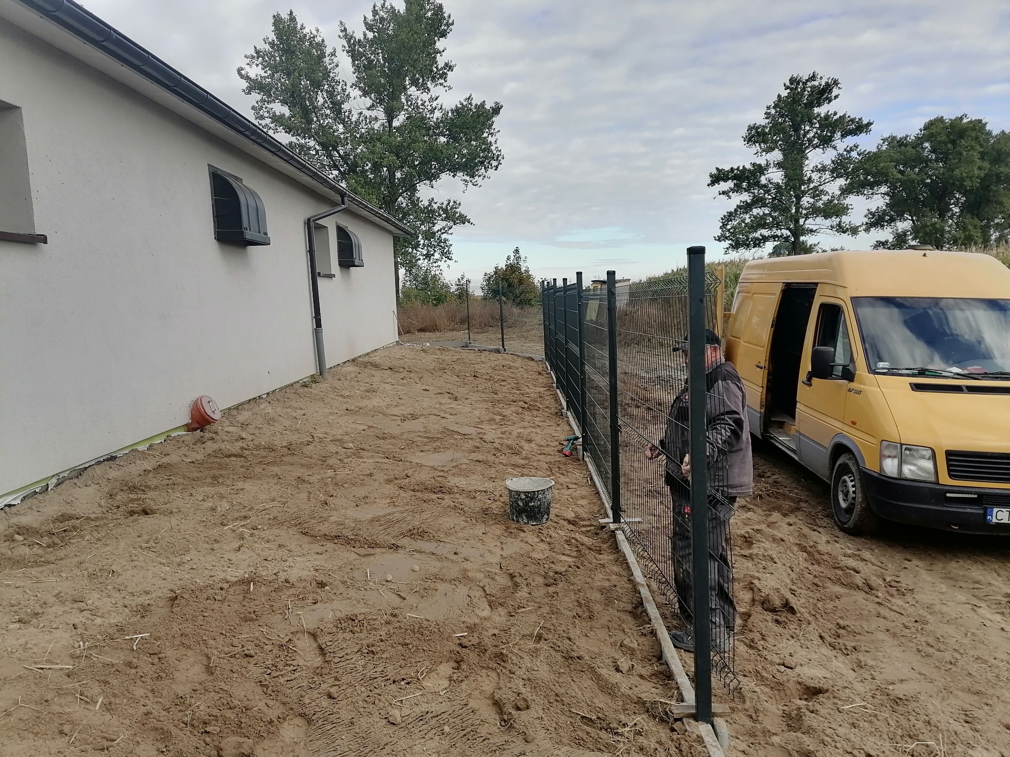 Montaż ogrodzenia panelowego przy budynku jednorodzinnym, widoczny pracownik w stroju roboczym, żółty bus z otwartymi drzwiami i teren przygotowany pod trawnik.