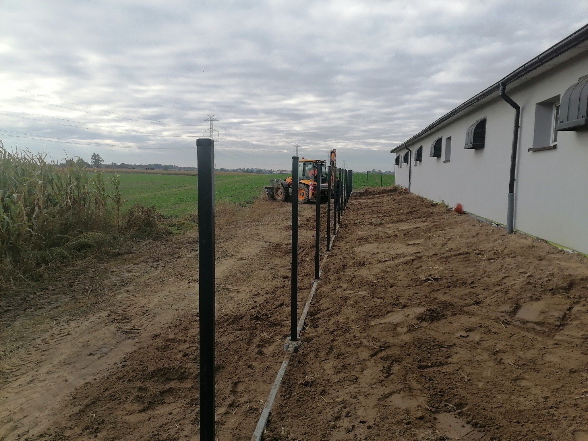 Montaż słupków ogrodzeniowych z użyciem traktora na tle pola i budynku gospodarczego w pochmurny dzień.